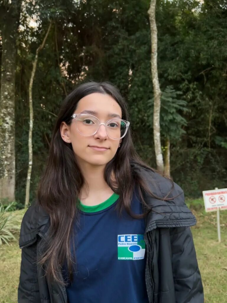 A imagem mostra uma jovem de cabelo preto, liso e longo, usando óculos de grau e sorrindo levemente para a câmera. Ela está de pé, atrás você percebe a natureza com muitas árvores. Ela veste uma jaqueta preta, uma camiseta azul com a logo representando a escola em que estuda CEEP (Centro Estadual de Educação Profissional Pedro Boaretto Neto). O fundo da imagem é um ambiente de natureza com árvores de diversos tipos.