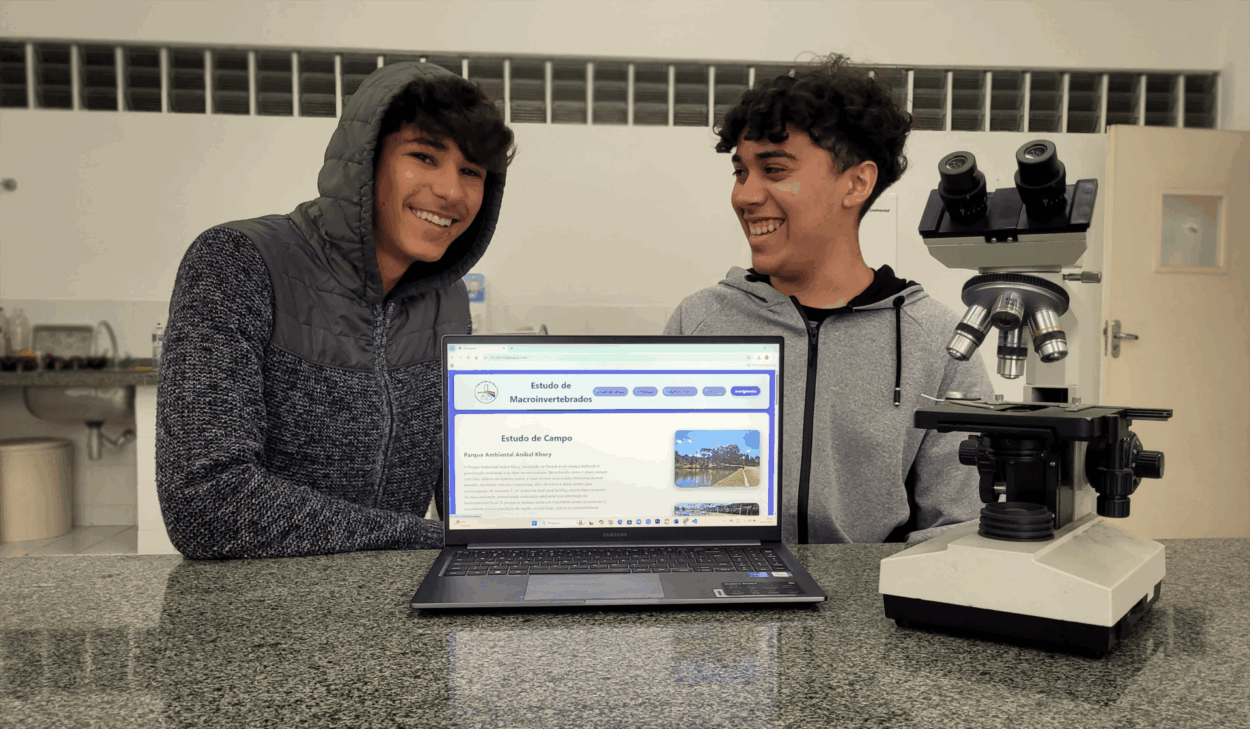 Dois estudantes estão sentados atrás de uma bancada de granito em um ambiente de laboratório escolar. Eles sorriem enquanto apresentam um notebook aberto com um site intitulado “Estudo de Macroinvertebrados”. O conteúdo na tela traz informações sobre o Parque Ambiental Anibal Khury e sua relação com estudos de campo. À direita do notebook, há um microscópio óptico, reforçando o caráter científico da atividade. Ambos os estudantes usam casacos. Ao fundo, é possível ver uma pia com materiais de laboratório e uma porta. A atividade faz parte de um projeto escolar de divulgação científica.
