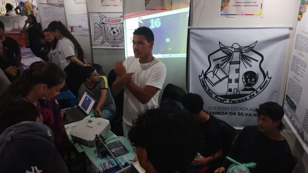 A imagem mostra um estande de feira científica ou educacional, com várias pessoas interagindo. No centro, um jovem de camiseta branca está falando e gesticulando, aparentemente explicando algo para o público. Sobre a mesa, há um projetor ligado e um notebook, com a projeção aparecendo na parede ao fundo. À direita, há um grande banner com o logotipo do “Colégio Estadual Professora Tereza da Silva Ramos”. Outras pessoas, incluindo visitantes e estudantes, observam e participam da atividade. O ambiente está movimentado, com materiais e cartazes afixados nas paredes do estande.