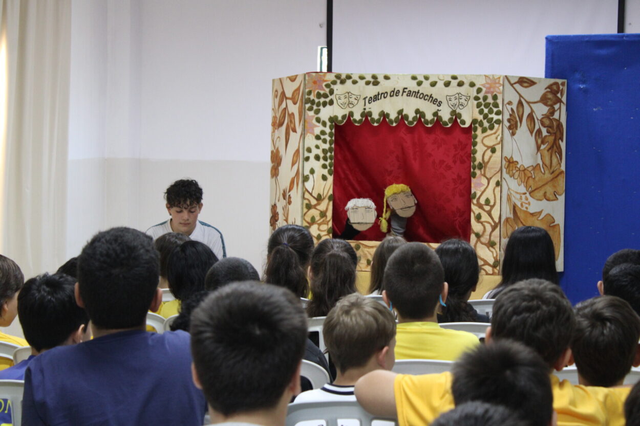 A foto mostra a apresentação de teatro de fantoches do clube Cabeceira do Rio Iapó para os estudantes do Colégio Municipal Dr. Vicente Machado. Ao fundo, vê-se um palco decorado com cortina vermelha e laterais com ilustrações de folhas e flores, onde dois fantoches estão sendo manipulados. À esquerda do palco, uma pessoa está sentada, concentrada, auxiliando na apresentação. Em primeiro plano, o público — composto por estudantes sentados em cadeiras plásticas — assiste atentamente à encenação. O ambiente é uma sala ampla, com paredes claras e boa iluminação.