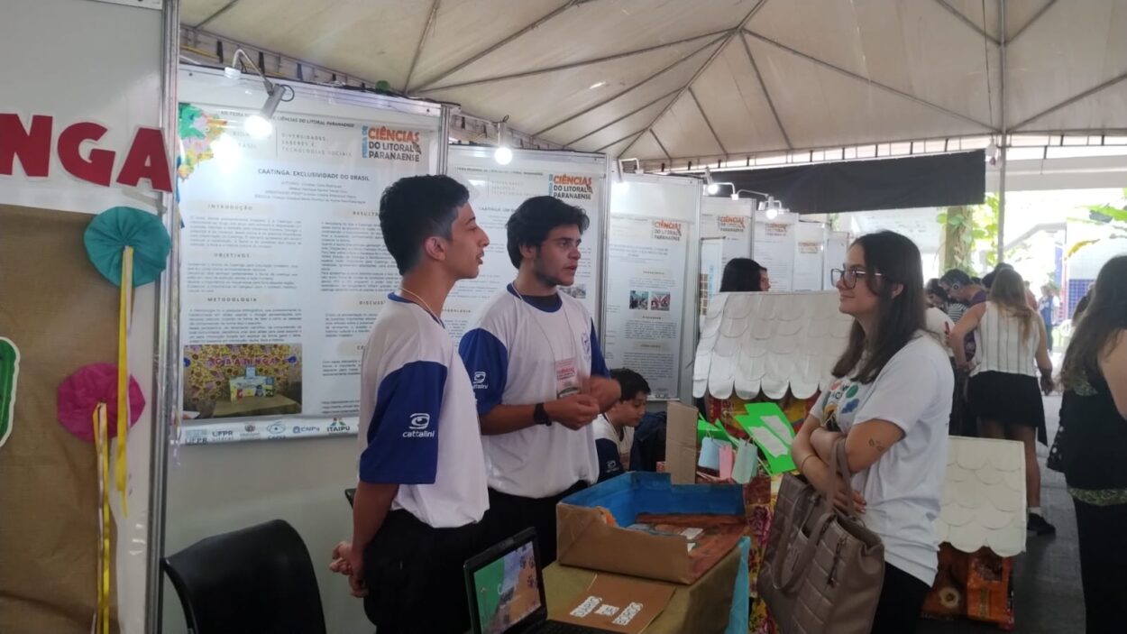 Na foto, dois estudantes, usando camisetas brancas com mangas azuis, apresentam seu projeto sobre a Caatinga para uma visitante, que escuta atentamente com os braços cruzados e segura uma bolsa bege. Ao fundo, vê-se um painel expositivo com o título “Caatinga: Exclusividade do Brasil” e outros trabalhos da feira de ciências, todos sob uma grande tenda. Sobre a mesa, há uma maquete colorida e um notebook aberto, compondo a exposição do grupo.