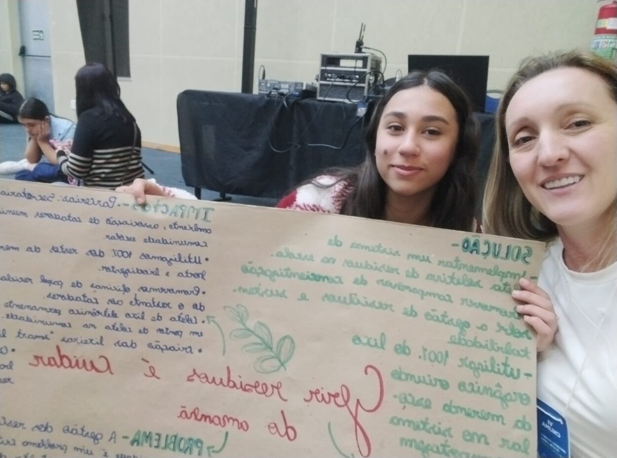 A imagem mostra duas pessoas: uma aluna, de cabelo comprido e liso, e uma professora, de cabelo loiro preso. Ambas sorriem, posando para a câmera. Elas seguram um cartaz com anotações coloridas. Trata-se das representantes do clube “Exploradores da Ciência”, do Colégio Estadual Campo Castelo Branco.