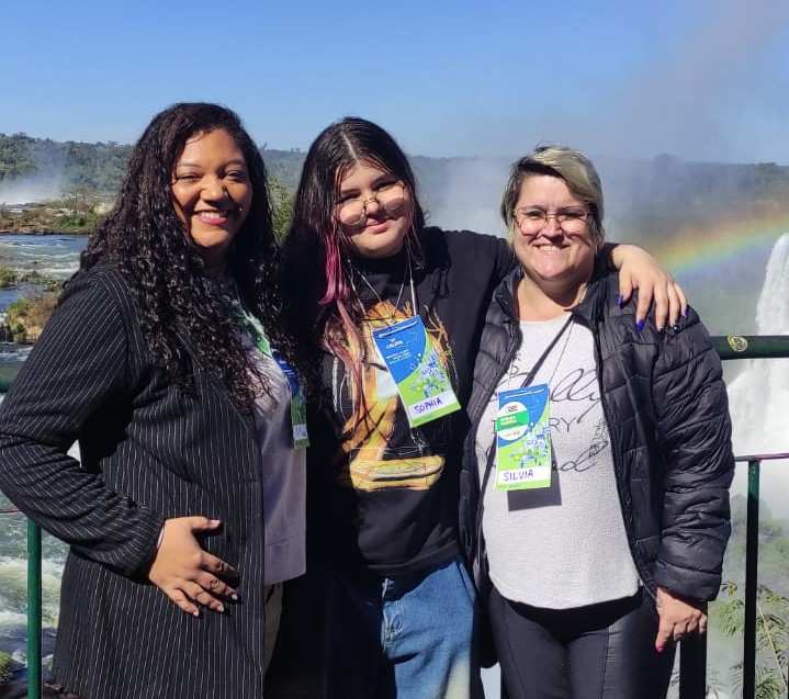 A imagem mostra três pessoas ao ar livre, sorrindo. As Cataratas do Iguaçu estão ao fundo e um arco-íris está visível. Trata-se das representantes do Clube Lumière, do Colégio Estadual Gustavo Dobrandino da Silva.