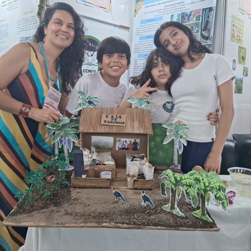 Na foto, uma professora e três estudantes sorriem ao lado de uma maquete intitulada “Casa de Farinha”. O trabalho é feito em papelão e decorado com figuras de plantas, animais e elementos da natureza, representando o ambiente de produção artesanal de farinha. Ao fundo, há painéis expositivos com informações científicas, indicando que a imagem foi registrada em um evento ou feira de ciências. Todos demonstram orgulho e entusiasmo pela apresentação do projeto.