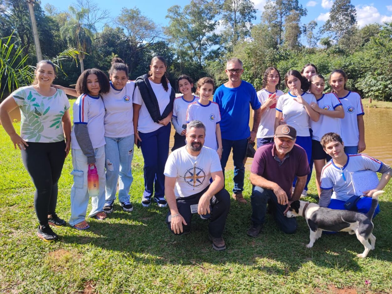 A imagem mostra um grupo de pessoas reunidas em um espaço ao ar livre, próximo a um lago e cercado por árvores. À esquerda, uma mulher de camiseta clara com estampa verde sorri para a câmera. Ao lado dela, vários estudantes em uniforme escolar branco com detalhes em azul e vermelho posam juntos. Dois homens adultos, um de camiseta azul e outro de camiseta branca com estampa, também participam da foto, assim como outro homem de boné e camiseta vinho que está agachado ao lado de um cachorro preto e branco.