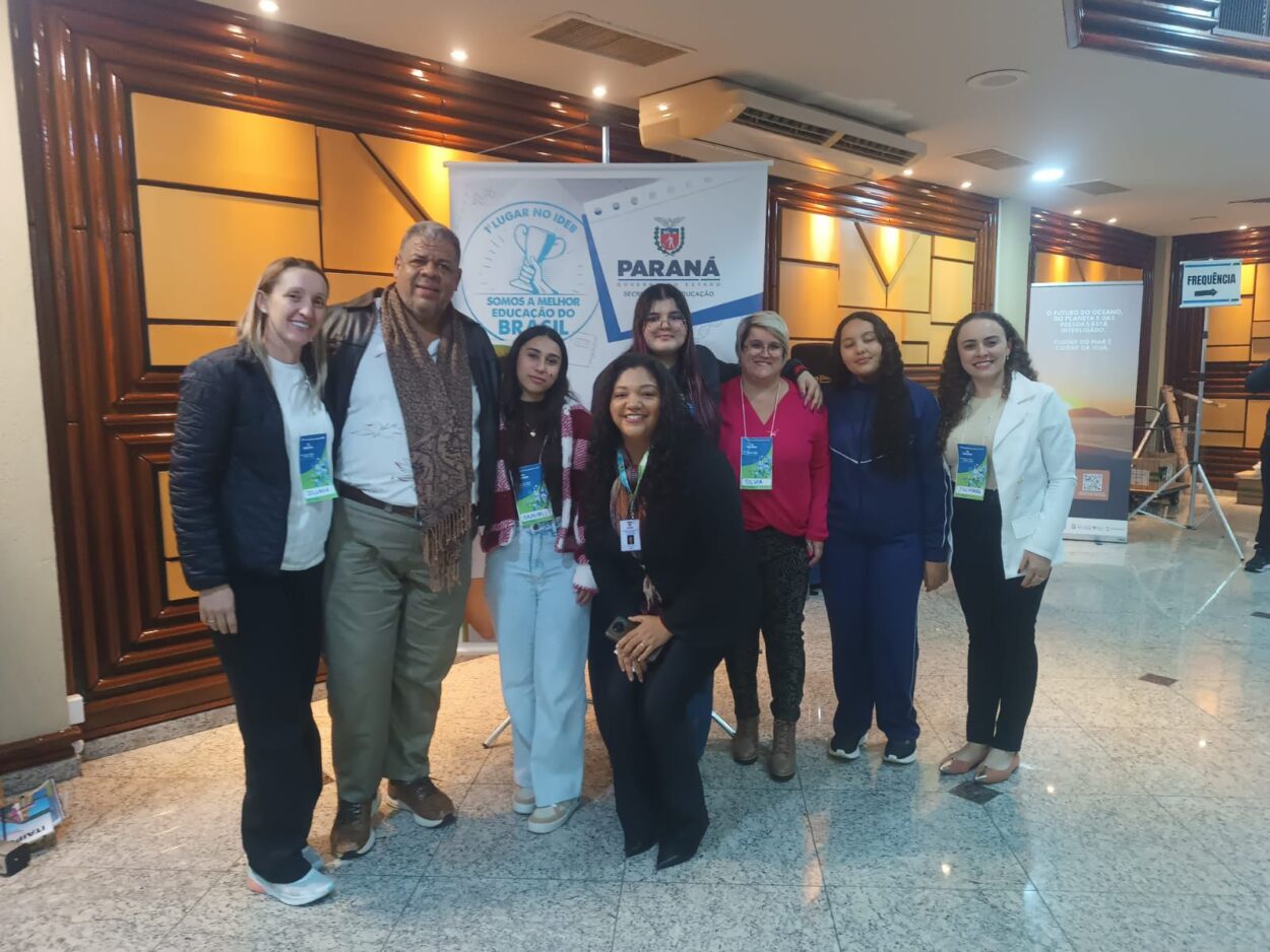 A imagem mostra um grupo de pessoas sorrindo para a câmera. Ao fundo, há um banner com o logotipo do Governo do Paraná. Trata-se de professores e alunos representantes dos clubes articulados pela Universidade Federal da Confederação Latino-Americana (UNILA). Eles estão posando durante a etapa estadual da VI Conferência Nacional Infantojuvenil pelo Meio Ambiente (CNIJMA).