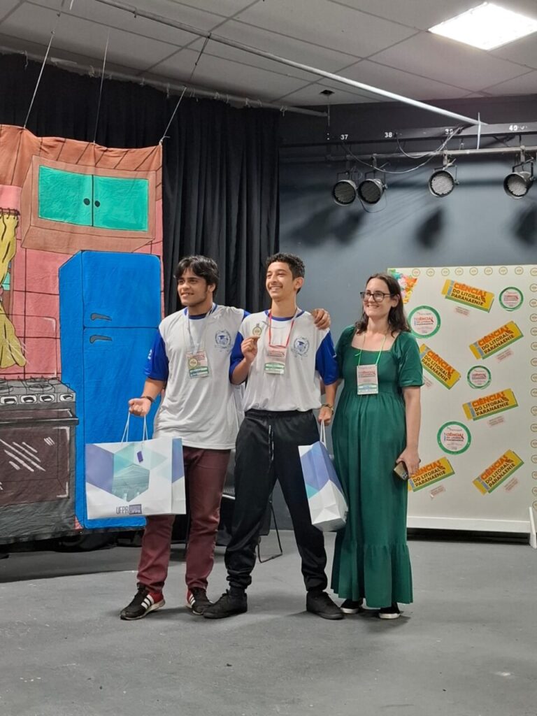 A imagem mostra dois estudantes e com a professora Andreia Petruy em um palco durante um evento. Os dois jovens, usando camiseta branca com mangas azuis, estão sorrindo, cada um segurando uma sacola de premiação. Um deles exibe também uma medalha no peito. Ao lado deles, a professora, de vestido verde, sorri enquanto posa para a foto. Ao fundo, há um painel colorido com a logo da "Ciência do Litoral Paranaense" e um cenário decorativo que imita uma cozinha desenhada. O clima é de celebração e reconhecimento.
