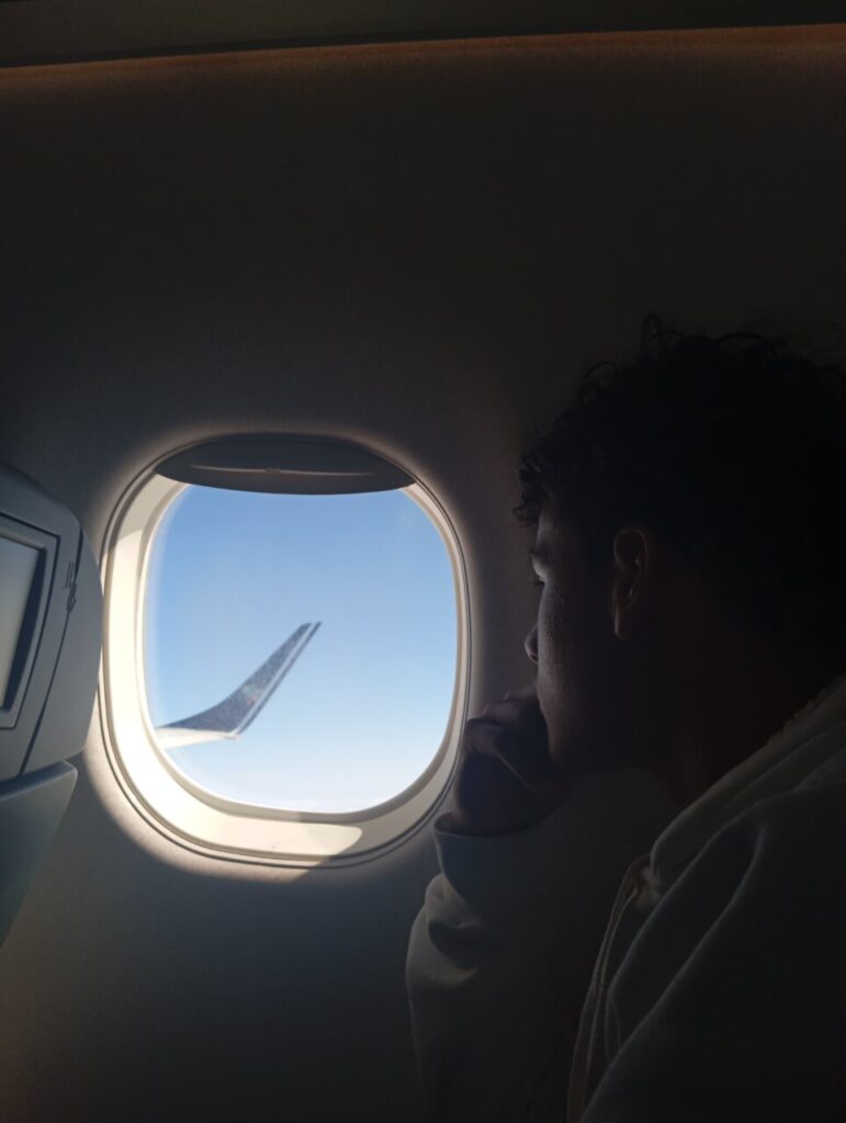 O estudante Renan está sentado ao lado da janela de um avião, olhando pensativamente para fora. A luz do sol ilumina parcialmente o interior da cabine, e a silhueta da pessoa aparece em contraste com a claridade vinda da janela. Do lado de fora, é possível ver parte da asa da aeronave recortando o céu azul. A cena transmite um momento de introspecção e tranquilidade durante o voo.