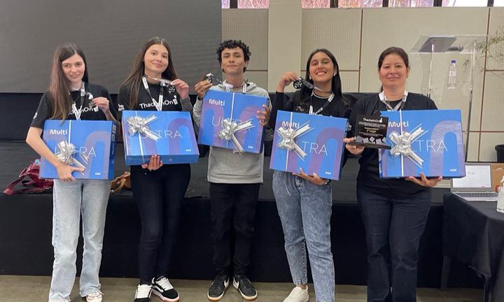 A imagem mostra um grupo de cinco pessoas (quatro jovens e uma mulher adulta) em pé, sorrindo e segurando prêmios. Todos usam camisetas pretas do evento "ThackathOn". Cada pessoa segura uma grande caixa azul com a palavra "ULTRA" em destaque e a marca "Multilaser", envolvida com um laço prateado, sugerindo que seja um notebook. Além disso, cada um segura um pequeno troféu preto.