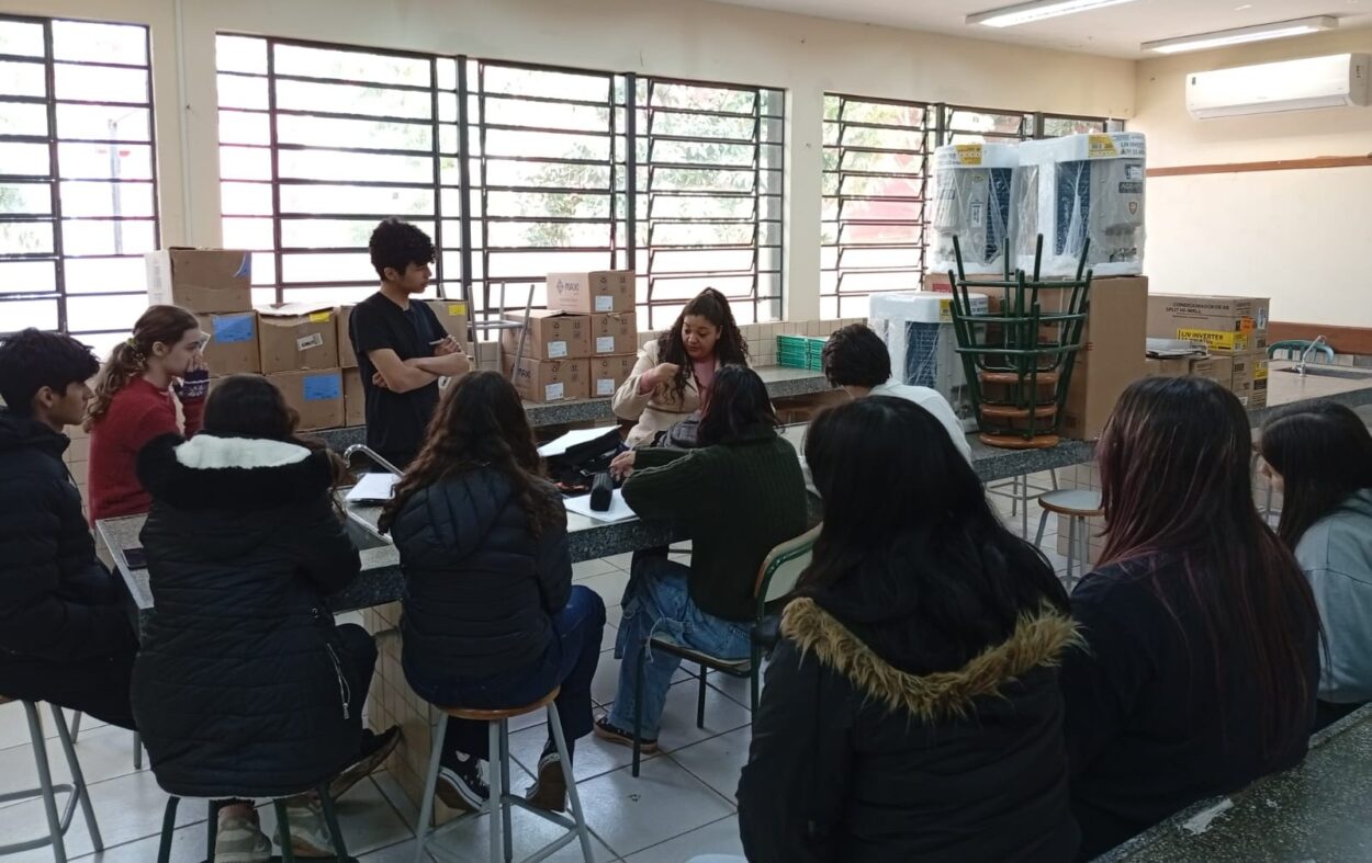 A foto mostra um grupo de estudantes reunidos em uma sala de aula com janelas amplas e muita luz natural. Eles estão em torno de uma bancada alta, parecida com as de laboratórios escolares. Uma jovem ao centro da imagem, sentada, está falando ou apresentando algo, enquanto os outros observam com atenção. Ao fundo, há várias caixas empilhadas e alguns equipamentos grandes ainda embalados. Também há cadeiras empilhadas de cabeça para baixo e um aparelho de ar-condicionado na parede. O ambiente é limpo, bem iluminado, com piso claro e janelas com grades.