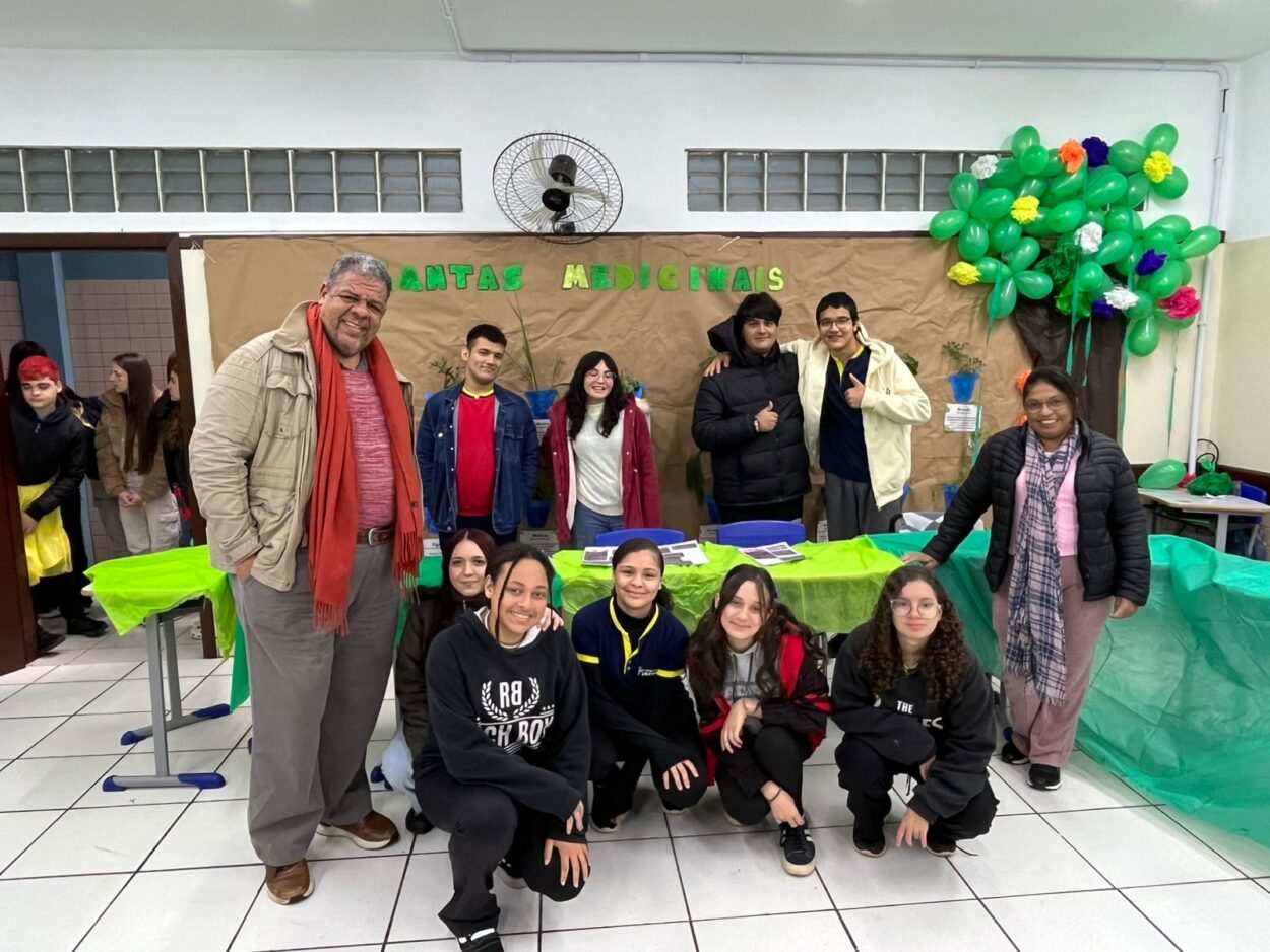 A imagem mostra um grupo de pessoas, entre estudantes e professores, posando sorridentes em uma sala decorada para uma apresentação escolar. Ao fundo, há um grande cartaz marrom com o título "PLANTAS MEDICINAIS" escrito com letras verdes e amarelas. No canto direito do cartaz, há uma árvore feita com bexigas verdes e flores coloridas. A mesa ao centro está coberta com um tecido verde vivo e há algumas plantas e materiais de apresentação sobre ela.