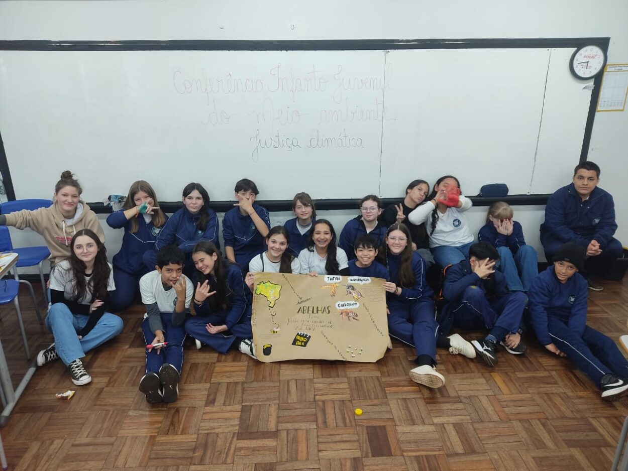 A imagem mostra um grupo de estudantes sentados no chão de uma sala de aula, todos sorrindo ou fazendo poses descontraídas. Há um quadro branco ao fundo, onde está escrito com caneta vermelha: “Conferência Infantojuvenil do Meio Ambiente – Justiça Climática”. No centro do grupo, alguns seguram um cartaz de papel pardo com o mapa da América do Sul desenhado. No cartaz está escrito “ABELHAS” em destaque, com figuras de abelhas e palavras relacionadas ao tema. O chão é de madeira, e há algumas cadeiras azuis ao lado esquerdo da imagem.
