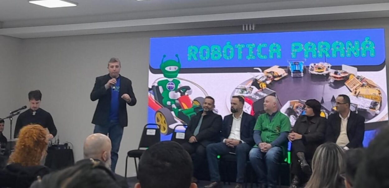 A imagem mostra um evento público em um auditório. No centro da cena, há um homem de pé segurando um microfone e falando ao público. Atrás dele, há um grande telão com a ilustração de um robô verde dirigindo um carro de corrida e várias miniaturas de robôs em uma arena. Acima da imagem no telão, lê-se “ROBÓTICA PARANÁ” em letras grandes e chamativas com estilo que remete a circuitos eletrônicos. À direita do orador, sete pessoas estão sentadas lado a lado em uma fileira de cadeiras, compondo uma mesa de autoridades ou convidados do evento. À esquerda da imagem, um homem de roupa preta está em pé, que opera equipamentos de som ou multimídia. Na frente, aparecem parcialmente várias pessoas da plateia, todas voltadas para o palco. O clima é de formalidade e interesse, típico de uma cerimônia ou apresentação oficial.