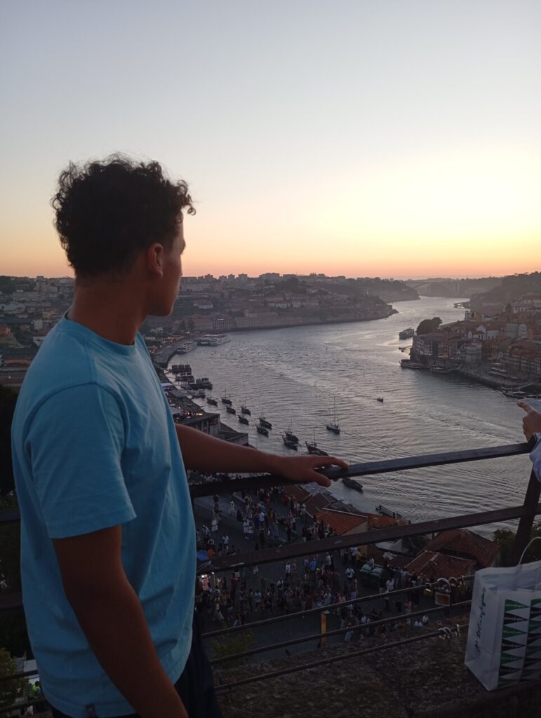 A imagem mostra um jovem de camiseta azul clara, visto de perfil, observando o pôr do sol sobre um rio. Ele está encostado em um corrimão, no alto de um mirante, com vista para a cidade e para diversas embarcações atracadas ou navegando. Ao fundo, é possível ver prédios, pontes e o céu em tons de laranja e rosa, refletindo na água. Na parte inferior da cena, há um grande grupo de pessoas reunidas, possivelmente turistas, aproveitando a vista. A atmosfera transmite tranquilidade e contemplação.