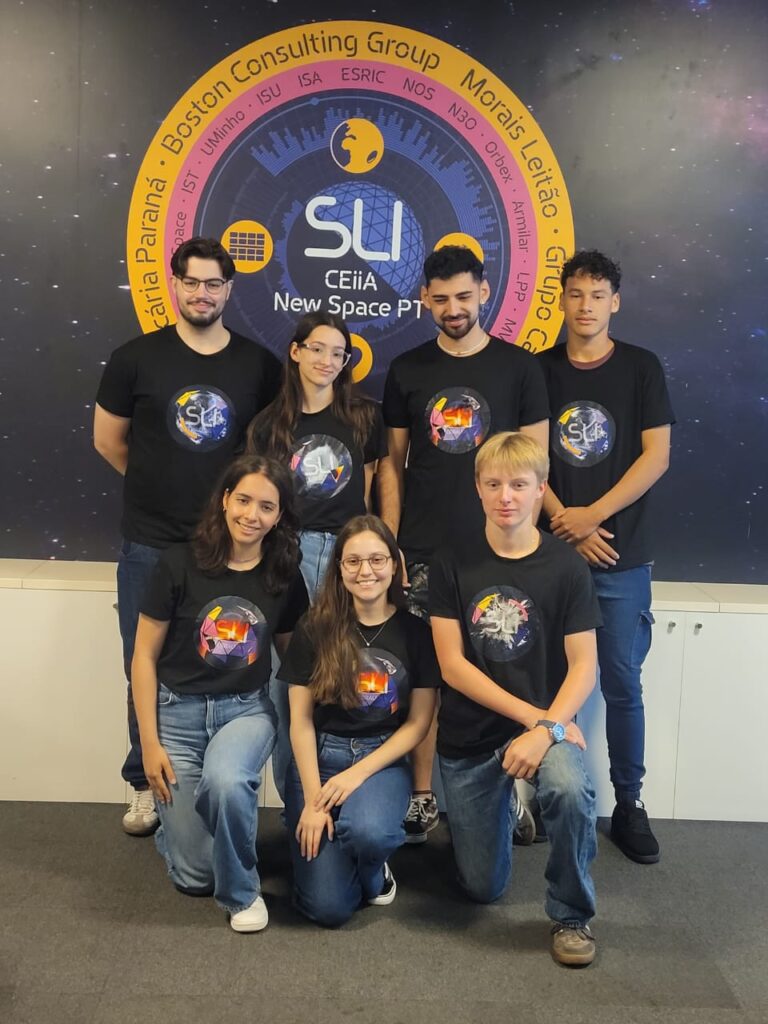 A imagem mostra um grupo de sete jovens posando para a foto em um ambiente interno. Eles vestem camisetas pretas com estampas coloridas relacionadas ao espaço e ao evento SLI (Sustainable Living Innovators), e usam calças jeans ou escuras. Ao fundo, há um grande painel com o logotipo do SLI e do CEiiA New Space PT, cercado por nomes de instituições e empresas parceiras. O grupo está disposto em duas fileiras: quatro pessoas em pé na parte de trás e três agachadas ou ajoelhadas na frente, todas sorrindo levemente para a câmera.