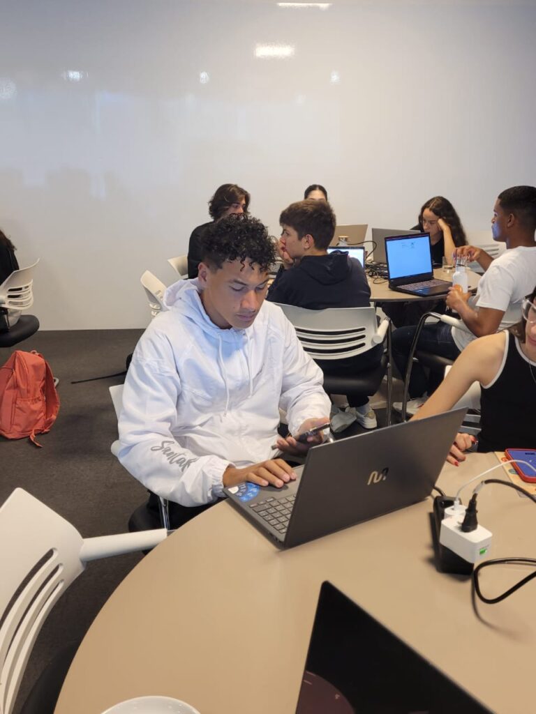 A imagem mostra um jovem usando um moletom branco sentado à frente de um notebook, enquanto segura um celular na mão. Ele está concentrado, trabalhando em um ambiente colaborativo, com outras pessoas ao fundo, também sentadas em mesas redondas e utilizando notebooks. O espaço é bem iluminado, com parede branca e cadeiras claras, transmitindo um clima de trabalho em grupo ou atividade acadêmica. Sobre a mesa, há cabos e um filtro de energia ligado a vários dispositivos.
