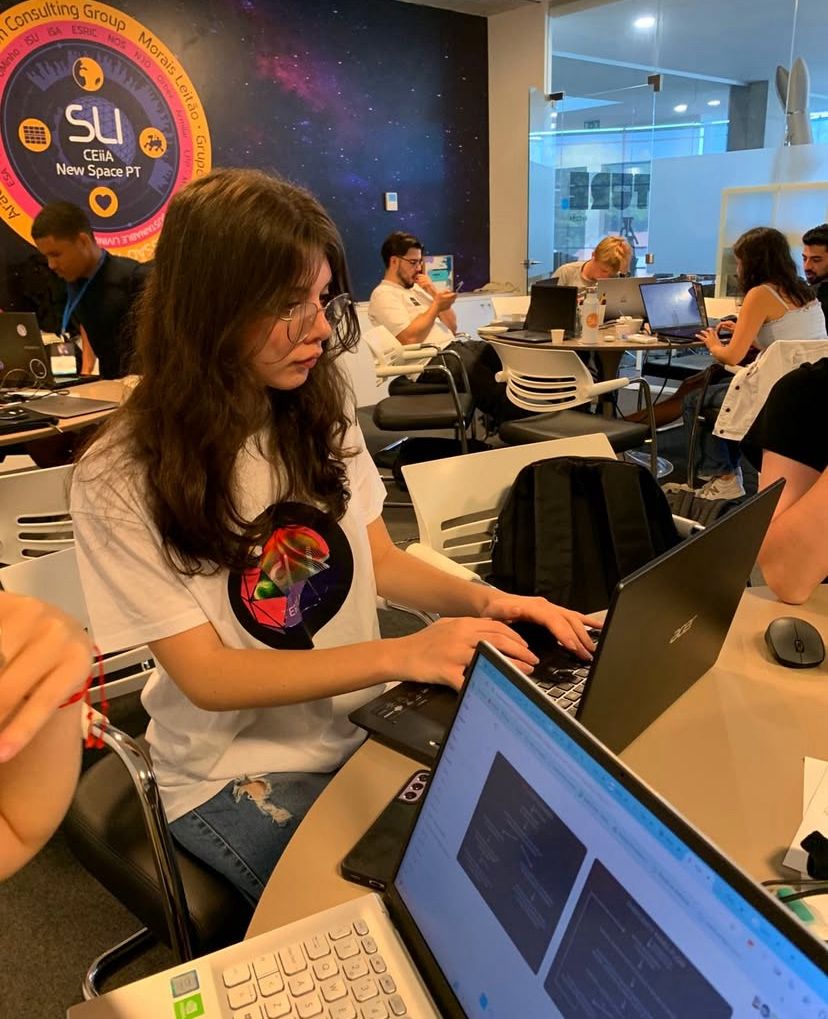A fotografia mostra uma jovem de cabelos longos e castanhos, usando óculos e camiseta branca com estampa colorida, sentada em uma mesa redonda e trabalhando em um notebook preto. À frente dela, há outro notebook aberto com uma tela clara. No fundo, outras pessoas também trabalham em computadores. Na parede, há um grande painel com o logotipo do programa “SLI – CEiiA New Space PT”, em um fundo que lembra o espaço sideral, com estrelas e planetas.