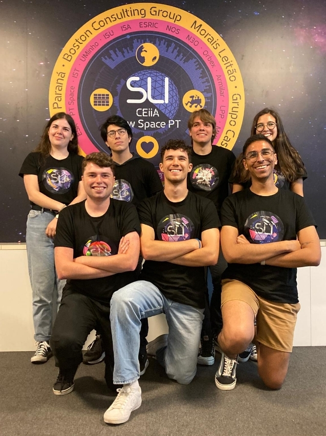 Na imagem, vemos um grupo de sete jovens posando juntos e sorridentes em frente a um painel com o logotipo do SLI – CEiiA New Space PT. Todos estão usando camisetas pretas com uma estampa colorida que remete ao espaço, reforçando o tema científico e tecnológico do projeto. Na parte de trás, o painel traz um círculo grande com nomes de instituições e empresas parceiras. O grupo está organizado em duas fileiras: quatro pessoas em pé atrás e três agachadas na frente, todos transmitindo energia positiva e espírito de equipe. O cenário sugere que eles participam de uma iniciativa voltada para ciência, tecnologia e exploração espacial