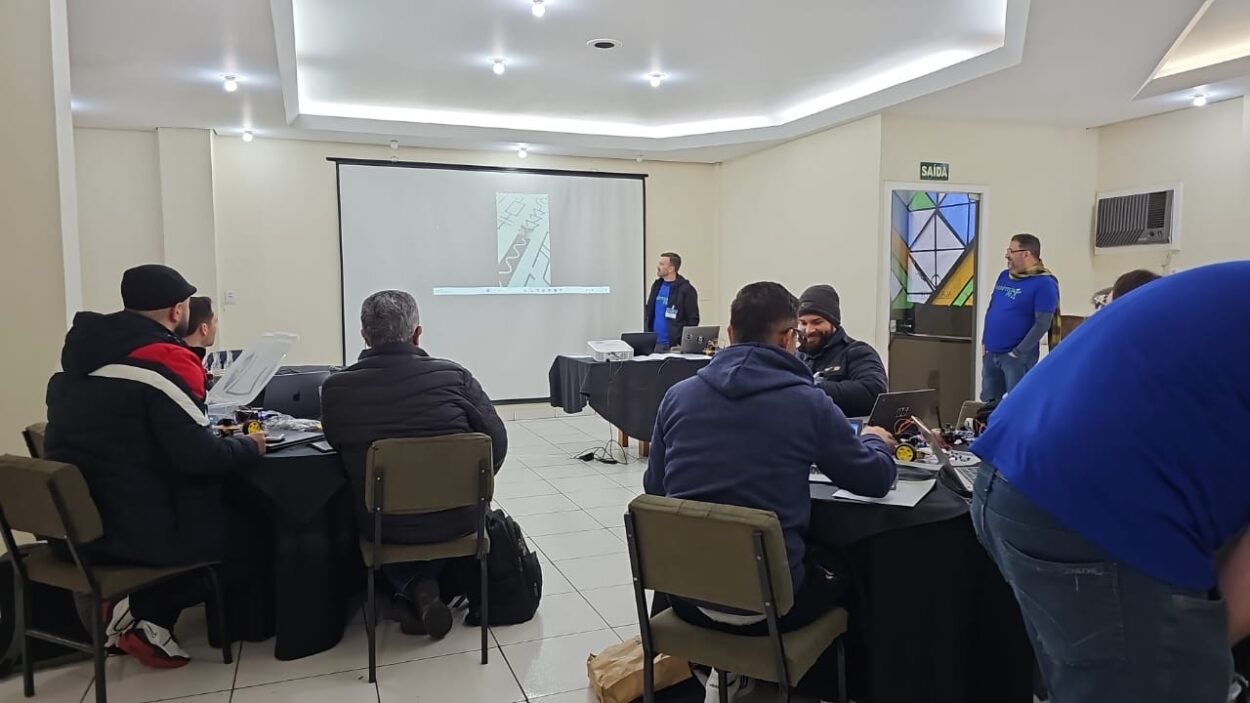 A galeria de fotos mostra momentos da oficina do 1ª Evento Presencial de Robótica 2025 em Curitiba, que acontece em ambientes fechados que alternam entre um auditório e uma sala com mesas, cadeiras, projetor e equipamentos eletrônicos. As fotos mostram professores construindo robôs e testando-os em uma pista no chão.