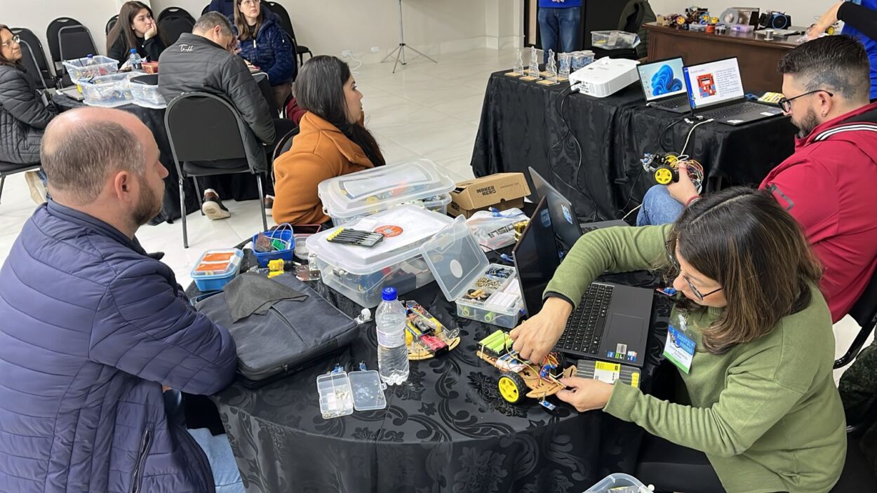 A galeria de fotos mostra momentos da oficina do 1ª Evento Presencial de Robótica 2025 em Curitiba, que acontece em ambientes fechados que alternam entre um auditório e uma sala com mesas, cadeiras, projetor e equipamentos eletrônicos. As fotos mostram professores construindo robôs e testando-os em uma pista no chão.