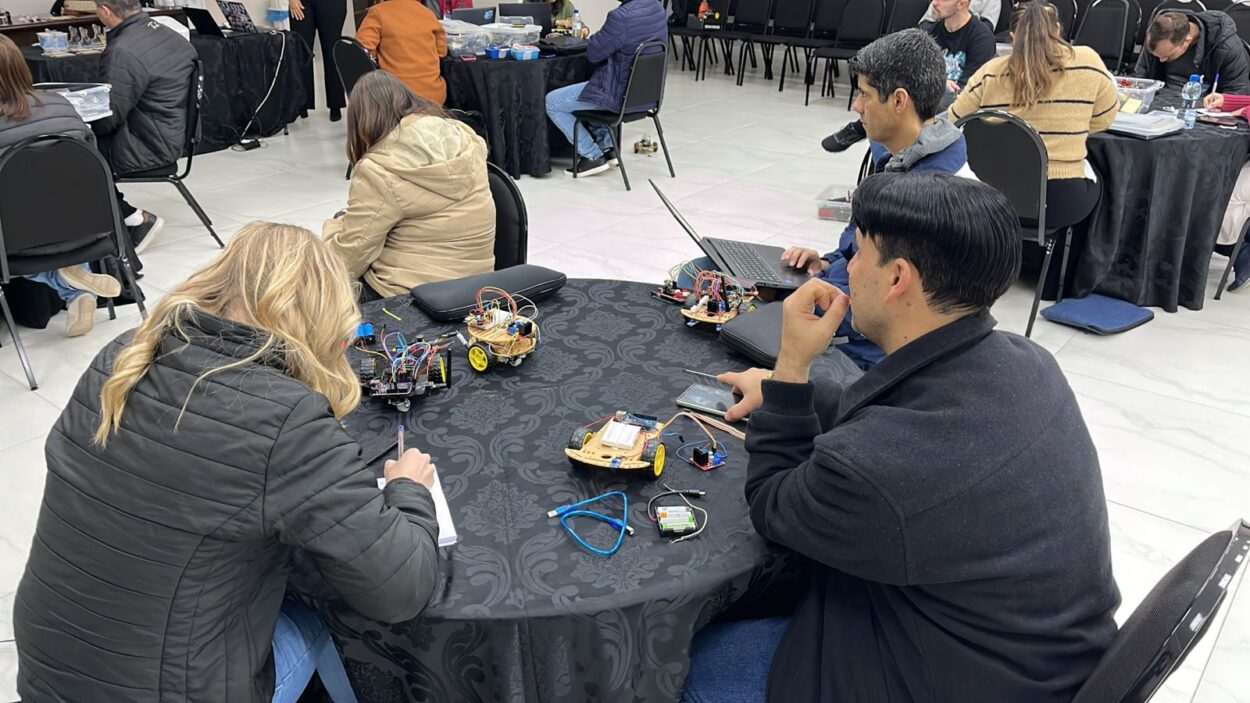 A galeria de fotos mostra momentos da oficina do 1ª Evento Presencial de Robótica 2025 em Curitiba, que acontece em ambientes fechados que alternam entre um auditório e uma sala com mesas, cadeiras, projetor e equipamentos eletrônicos. As fotos mostram professores construindo robôs e testando-os em uma pista no chão.