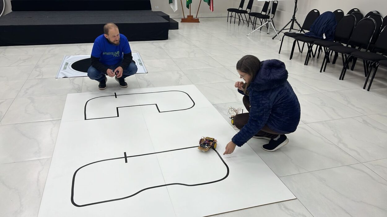A galeria de fotos mostra momentos da oficina do 1ª Evento Presencial de Robótica 2025 em Curitiba, que acontece em ambientes fechados que alternam entre um auditório e uma sala com mesas, cadeiras, projetor e equipamentos eletrônicos. As fotos mostram professores construindo robôs e testando-os em uma pista no chão.