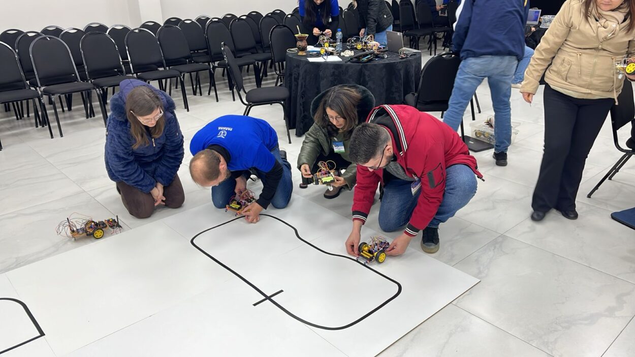 A galeria de fotos mostra momentos da oficina do 1ª Evento Presencial de Robótica 2025 em Curitiba, que acontece em ambientes fechados que alternam entre um auditório e uma sala com mesas, cadeiras, projetor e equipamentos eletrônicos. As fotos mostram professores construindo robôs e testando-os em uma pista no chão.
