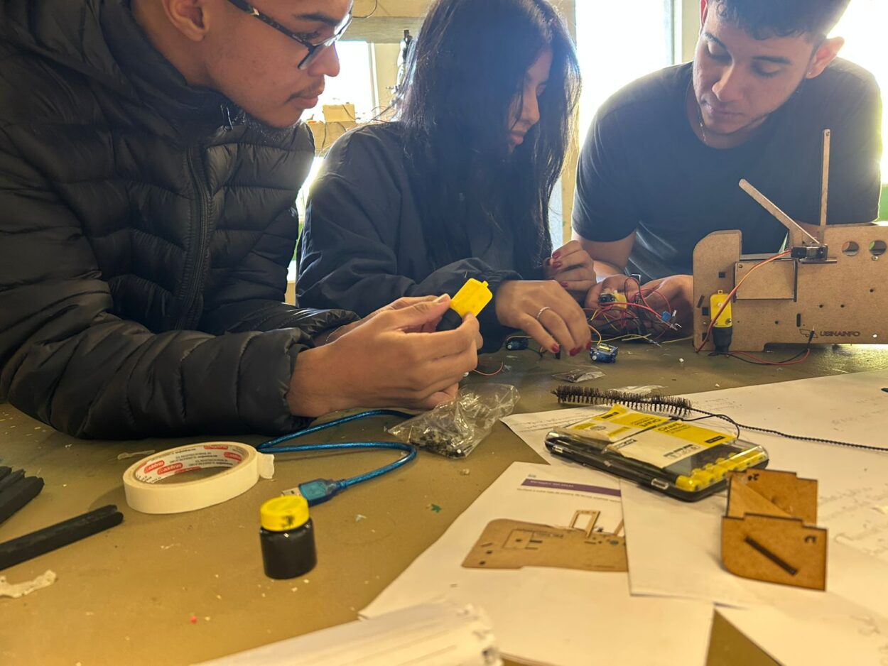 A galeria de fotos mostra o processo de construção de 3 projetos feitos por alunos do ensino médio e fundamental em um ambiente escolar. O primeiro projeto é de uma maquete de uma fazenda sustentável, o segundo projeto está no início de construção e mostra componentes eletrônicos utilizados em projetos de robótica ou automação para a produção de uma bengala inteligente para um usuário cego; e o terceiro projeto também está no início de produção e trata-se de um protótipo de esteira para separação de alimentos.