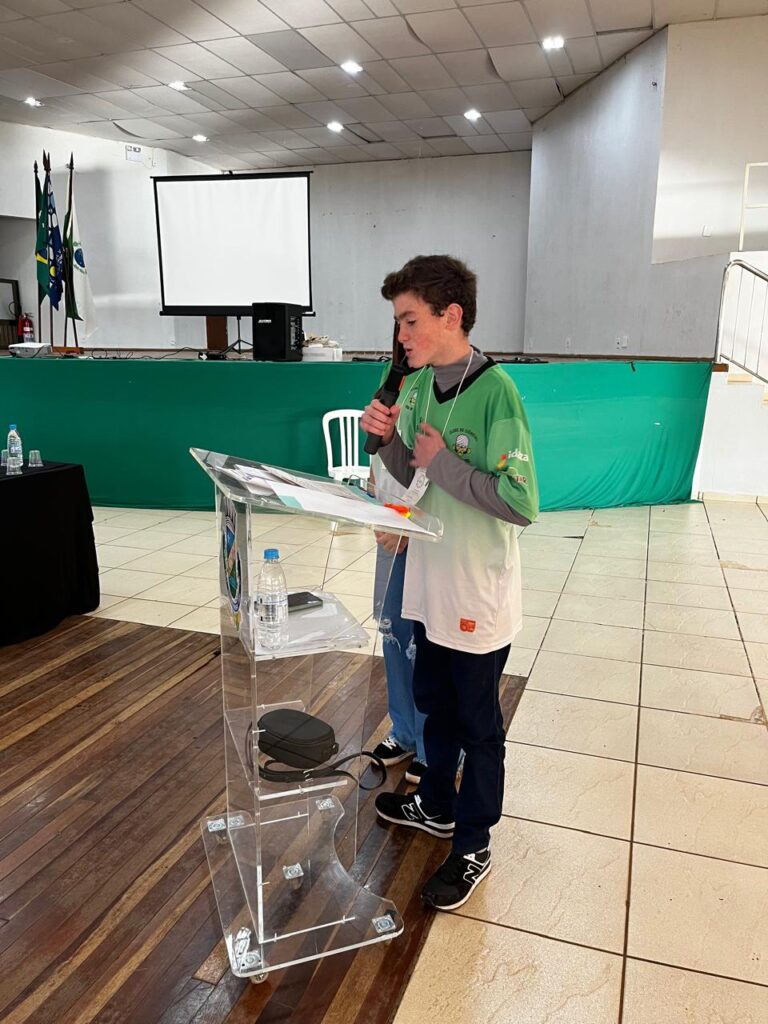 A imagem de um menino segurando o microfone, mostra seu diálogo com o público ao apresentar seu projeto, na frente dos demais envolvidos, alunos e professores.