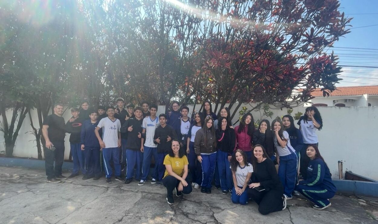 A foto é do Clube de Ciências Jovens Cientistas Kennedy. A imagem mostra uma turma de estudantes posando para uma foto acompanhada por três adultos, sendo um professor coordenador e duas Bolsistas Técnicas de Nível Superior - Pedagógicas da Rede de Clubes Paraná Faz Ciência em um dia de visita ao clube. Eles estão reunidos em um pátio externo escolar. Ao fundo, há árvores de folhas vermelhas e uma parede branca que cerca o local. A maioria dos alunos veste uniforme escolar. O grupo está posicionado em frente às árvores, formando duas fileiras: a maior parte em pé e alguns agachados ou ajoelhados à frente. Todos parecem estar posando para a foto coletiva, muitos sorrindo e fazendo gestos com as mãos, transmitindo um clima de descontração e união.