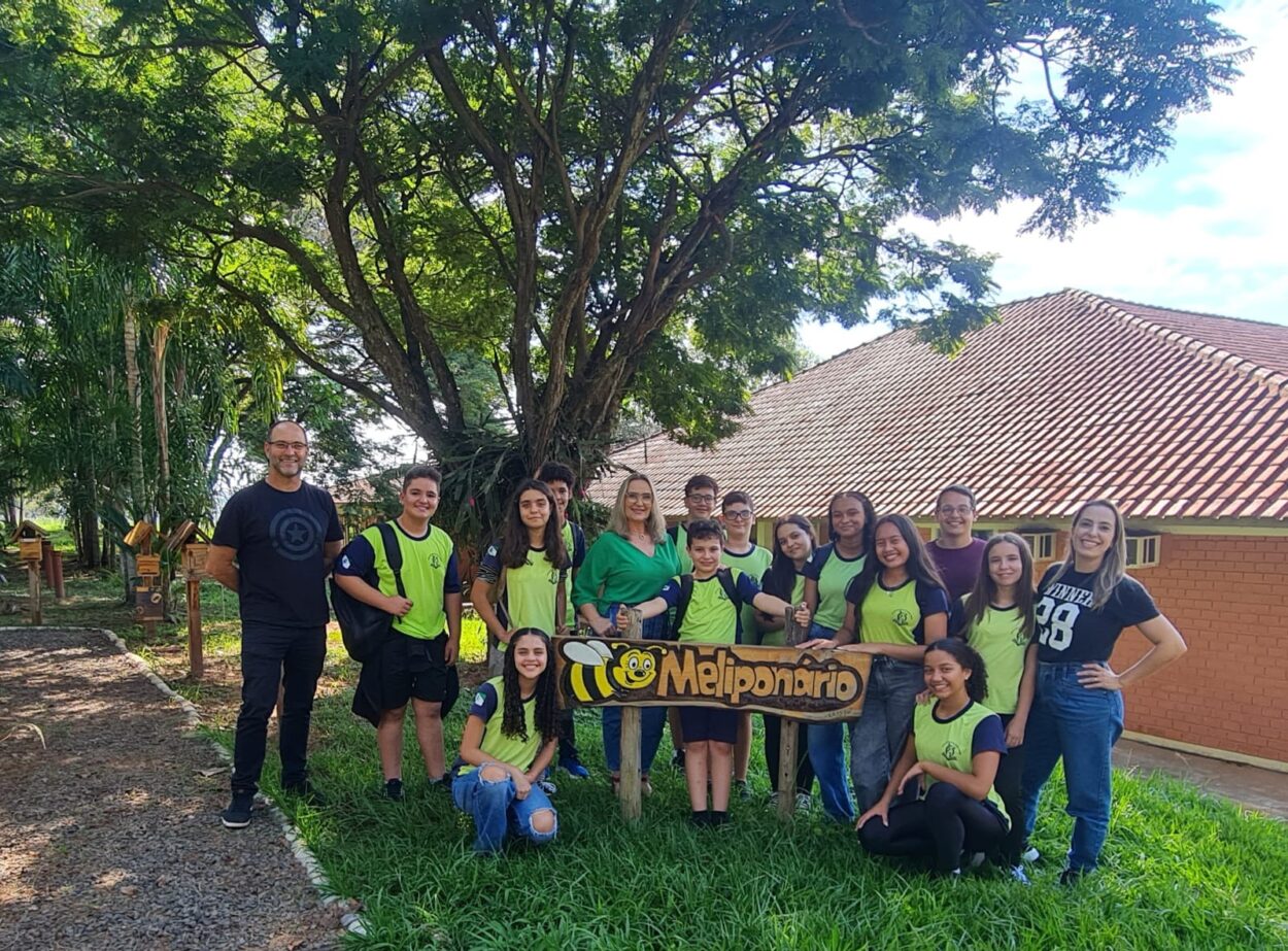 A primeira imagem é do Clube Experimentando a Ciência: Descobrindo cores e novos sabores. A imagem mostra um grupo de estudantes e professores posando para uma foto ao ar livre, em frente a uma placa de madeira com o desenho de uma abelha e a palavra “Meliponário”. O local é arborizado, com gramado verde, árvores grandes e, ao fundo, um prédio de tijolos com telhado de telhas vermelhas. A maioria dos alunos veste uniforme escolar, alguns usam mochilas. Estão sorridentes e organizados ao redor da placa. A segunda imagem é do Clube Ilha da Ciência. A imagem mostra um grupo de estudantes e duas mulheres posando em um ambiente ao ar livre, com árvores, arbustos e canteiros de horta ao fundo. A maioria dos alunos veste camisetas com logotipo escolar, e alguns carregam mochilas. O grupo está sorridente e reunido de forma descontraída, alguns em pé e outros agachados na frente.