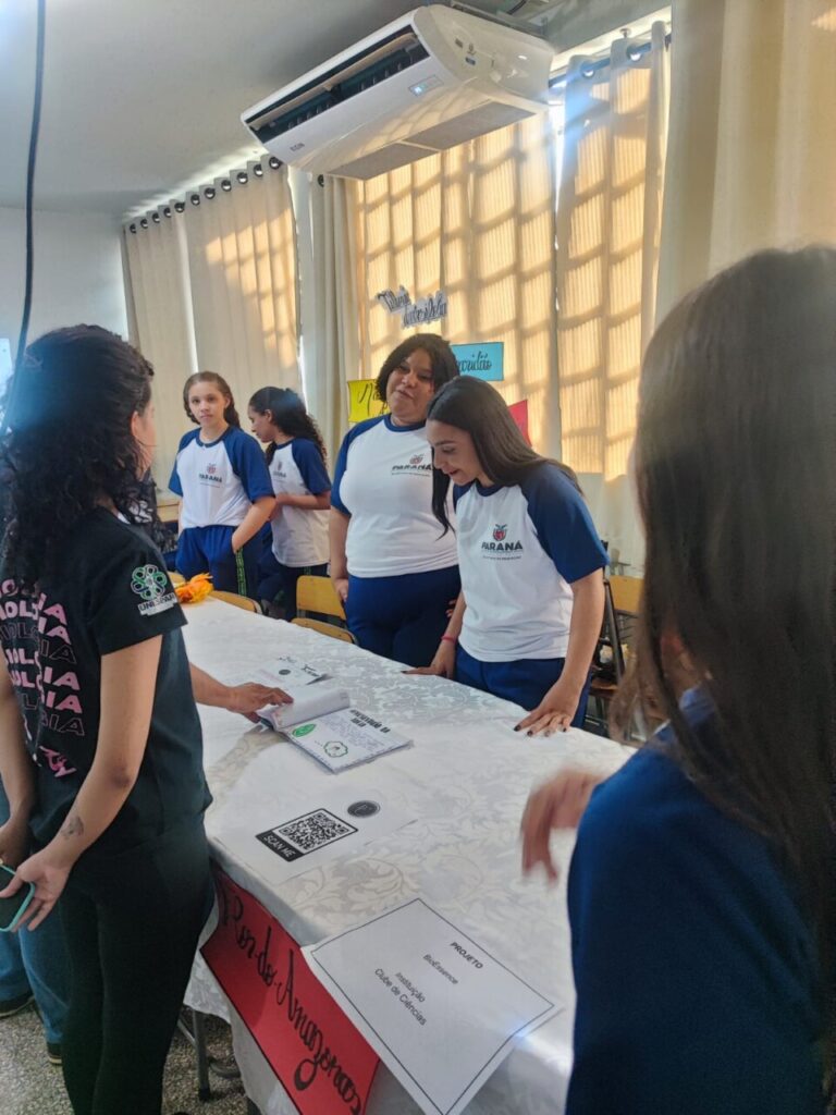 Na foto, estudantes apresentam um projeto durante a 1ª Feira de Ciências da Unespar – Campus Paranavaí. Duas alunas, usando uniforme escolar, estão atrás de uma mesa coberta com toalha branca, onde se vêem materiais do trabalho exposto, incluindo cartazes, panfletos e um QR Code para consulta digital.