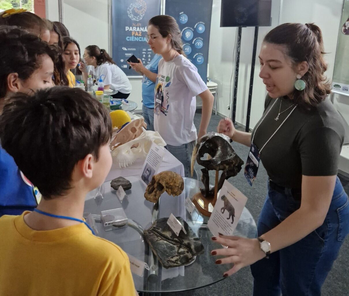 A imagem mostra um estande de divulgação científica. Uma expositora conversa com um grupo de crianças, explicando réplicas de fósseis e crânios,dispostos sobre uma mesa com placas informativas. Ao fundo, há um banner com os dizeres “NAPI PARANÁ FAZ CIÊNCIA”, indicando que o evento é uma iniciativa de promoção científica no estado do Paraná, Brasil. Outras pessoas, incluindo organizadores e visitantes, também estão presentes ao fundo, interagindo com diferentes materiais.