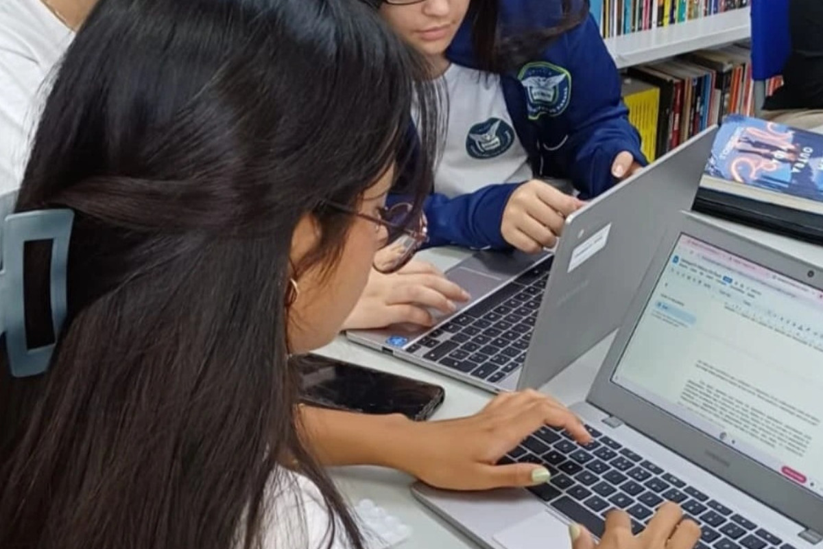 A imagem mostra alunas sentadas lado a lado em frente a notebooks prateados, digitando e trabalhando juntas. Estão em uma biblioteca, pois há prateleiras com livros ao fundo. Uma das alunas aparece em primeiro plano, com os cabelos longos e presos com uma presilha. Elas estão finalizando os projetos que apresentarão no evento.