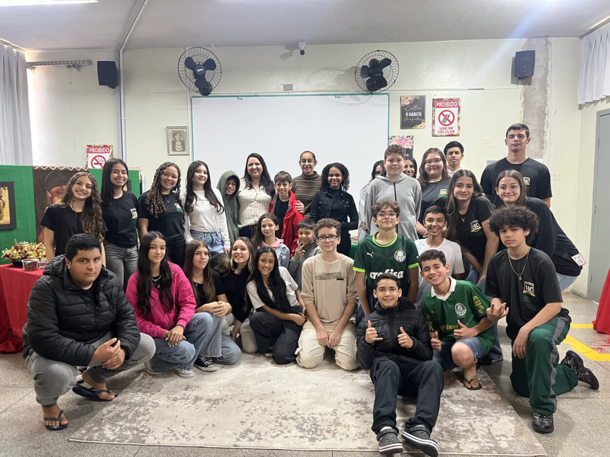 A imagem é do clube ‘Tenda Cultura Viva’ do Colégio Estadual Professora Hilda Trautwein Kamal de Umuarama. Na fotografia é possível ver um grupo grande de pessoas, formado por adolescentes e um adulto, posando para uma foto em uma sala. Eles estão organizados em duas fileiras: alguns sentados ou agachados na frente e outros em pé atrás. A sala tem paredes claras, ventiladores fixados na parede e um quadro branco ao fundo. Há cartazes e avisos na parede. À esquerda, há uma mesa com uma toalha vermelha e alguns objetos decorativos. Todos parecem estar sorrindo ou com expressões amigáveis.