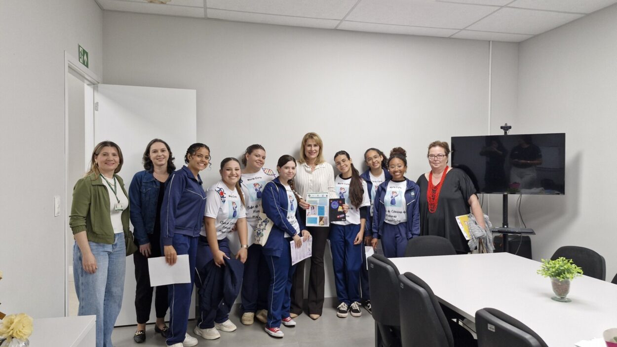Algumas estudantes estão sentadas ao redor de uma mesa redonda branca. Elas usam o mesmo uniforme de Marie Curie e seguram papéis coloridos. Na mesa, há folhetos impressos em tons de roxo e branco, com textos sobre ciência e empoderamento feminino.