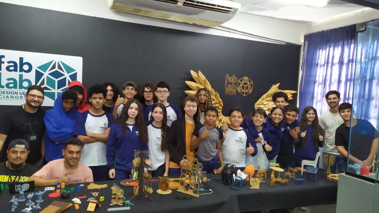 A imagem mostra um grupo de estudantes e adultos posando para uma foto em um ambiente interno, em frente a uma parede preta com os logotipos “fab lab” e elementos gráficos dourados que lembram asas. Na frente do grupo, há uma mesa com várias peças coloridas, miniaturas e objetos criativos, provavelmente produzidos em impressoras 3D ou cortadoras a laser, como chaveiros, estruturas geométricas e pequenos móveis. A maioria dos estudantes vestem camisetas escolares.