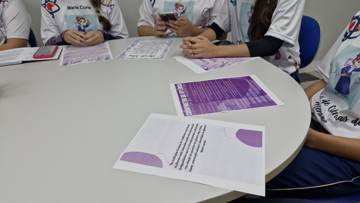 Algumas estudantes estão sentadas ao redor de uma mesa redonda branca. Elas usam o mesmo uniforme de Marie Curie e seguram papéis coloridos. Na mesa, há folhetos impressos em tons de roxo e branco, com textos sobre ciência e empoderamento feminino.