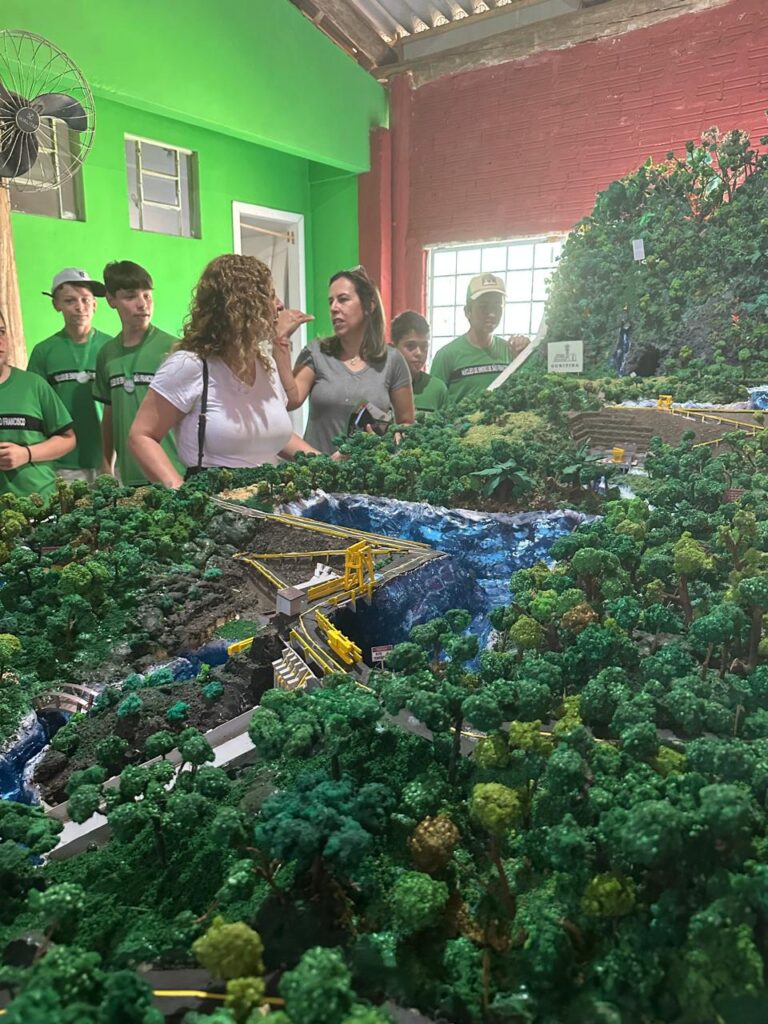Na foto, pessoas observam a maquete da usina hidrelétrica, que é composta por mini árvores e um rio de água corrente