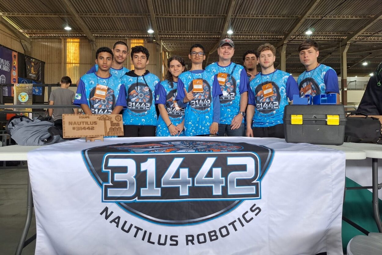 Um grupo de nove integrantes da equipe de robótica Nautilus Robotics #31442 está reunido em um espaço coberto de competição. Eles vestem uniformes azuis estampados com o logotipo da equipe e o número de identificação, posando lado a lado atrás de uma mesa de apoio. Sobre a mesa, há uma caixa de ferramentas preta com travas amarelas, uma placa de madeira gravada com o nome da equipe e uma grande faixa branca com o número 31442 em destaque. Ao fundo, banners coloridos de outros times e a estrutura metálica do ginásio completam o cenário, reforçando o ambiente de torneio de robótica.