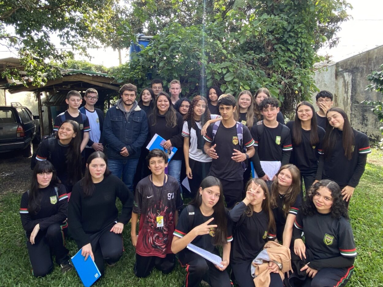 Fotografia tirada ao ar livre, durante o dia, com cerca de 30 pessoas posando para a câmera. A maioria é de adolescentes usando uniforme escolar preto com detalhes em verde, vermelho e branco nas mangas e o brasão da escola no peito. Eles estão agrupados em um gramado, cercados por árvores e vegetação ao fundo. Algumas pessoas estão sorrindo, outras fazendo gestos com as mãos, como sinais de paz e vitória. Duas pessoas adultas, ao centro da imagem, estão vestidas com roupas casuais e seguram pastas azuis. Ao fundo, vê-se uma cobertura com carros estacionados e um muro coberto por vegetação. A luz do sol ilumina suavemente o ambiente.