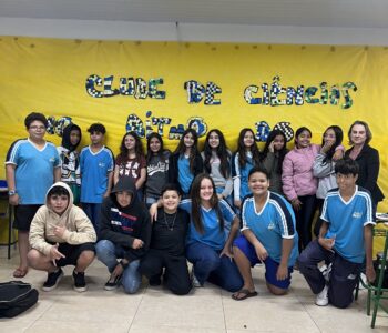 Foto dos clubistas e da professora coordenadora do clube No Ritmo do Conhecimento, em frente ao mural feito pelos estudantes (Foto/ Ana Carolina A. Barbosa)
