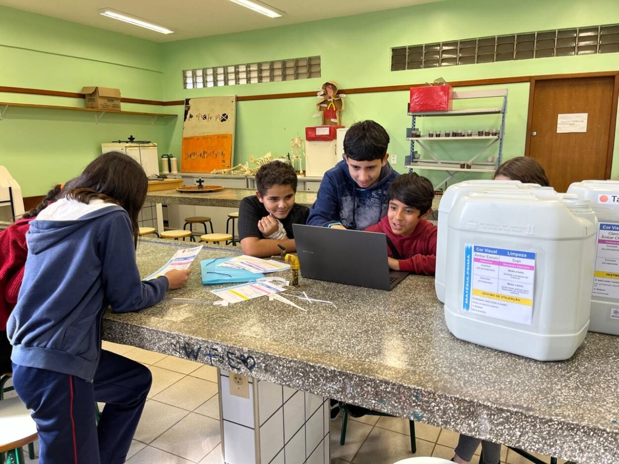 Na foto, alunos estão em um laboratório de ciências, trabalhando em uma bancada. Um dos estudantes toma notas enquanto outros visualizam algo na tela de um notebook.