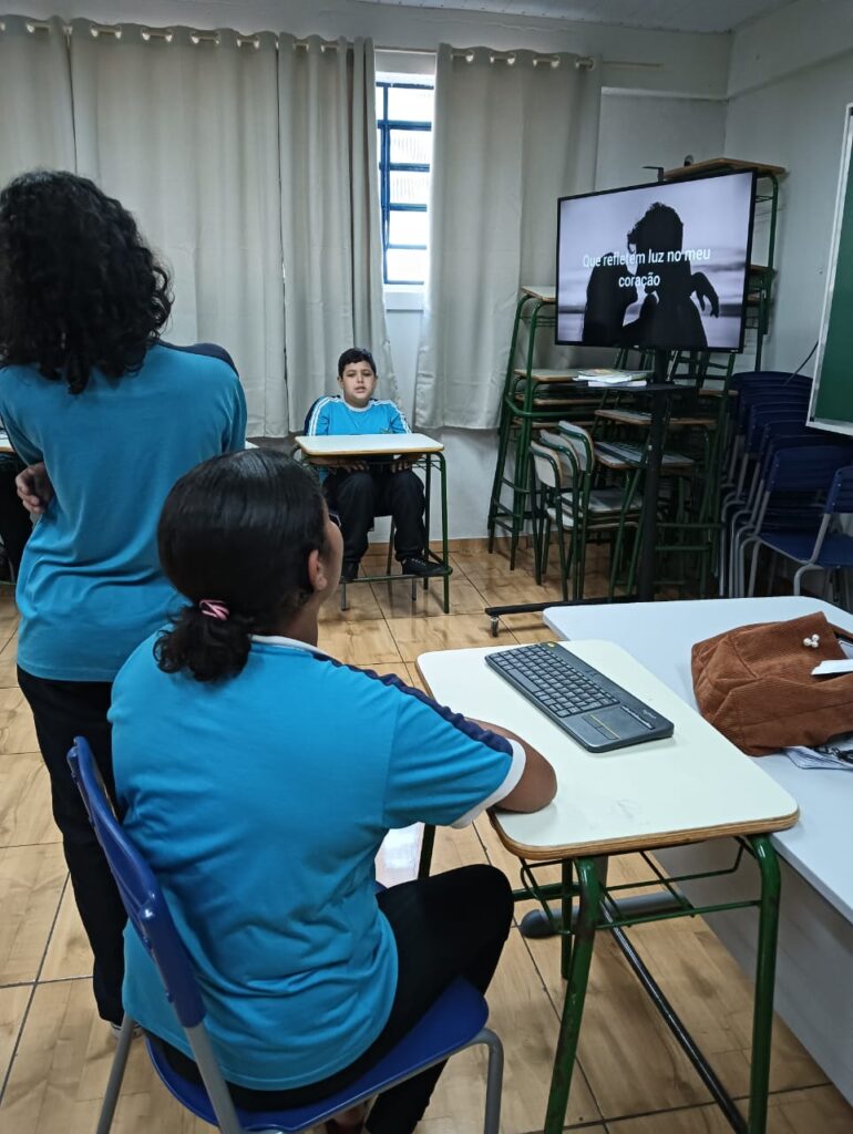 A imagem mostra uma sala de aula onde ocorre uma atividade educativa multimídia. Estudantes, vestindo camisetas azuis do uniforme escolar, participam da dinâmica. Um dos alunos está sentado de frente para uma televisão que exibe a frase: "Que refletem luz no meu coração", sugerindo que a turma esteja ouvindo uma música. No centro da imagem, uma aluna está sentada à mesa com um teclado sem fio à sua frente, indicando o uso de tecnologia durante a atividade. Ao fundo, outro aluno está sentado sozinho diante da janela com cortinas claras. A sala possui piso de madeira clara, carteiras organizadas e cadeiras empilhadas no canto, demonstrando um ambiente preparado para diferentes atividades. A cena transmite um momento de concentração e envolvimento dos alunos.