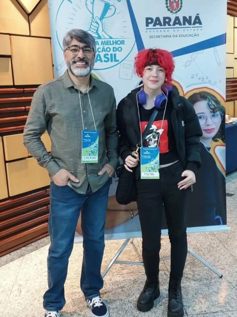 Duas pessoas estão em pé e sorrindo para a foto. À esquerda, um homem de cabelos e barba grisalhos, usa óculos, camisa cinza de manga comprida e calça jeans azul. À direita, uma jovem com cabelo curto e vermelho, veste jaqueta preta, calça preta e botas pretas. Ela tem um fone de ouvido roxo no pescoço e uma camiseta preta com estampa vermelha. Ambos usam crachás de identificação pendurados no pescoço. Ao fundo, há um banner da Secretaria da Educação do Paraná com frases e ilustrações coloridas.