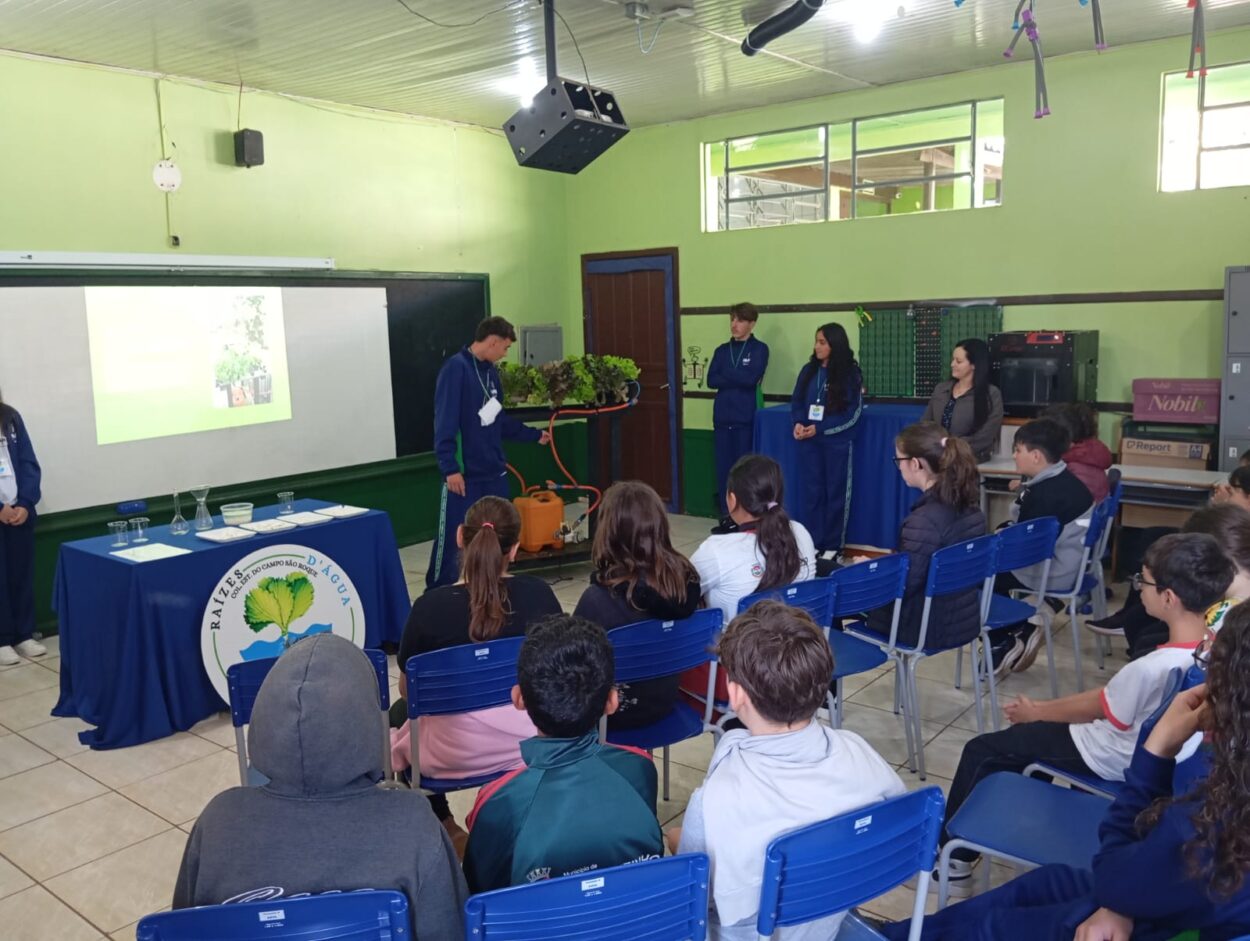 A foto mostra clubistas do Raízes D’Água, que participarão do 5º Paraná Faz Ciência. Eles estão apresentando seu projeto em uma sala de aula, para outros alunos, que permanecem atentos. Ao seu lado, seu protótipo de horta hidropônica. (Foto/Talula)