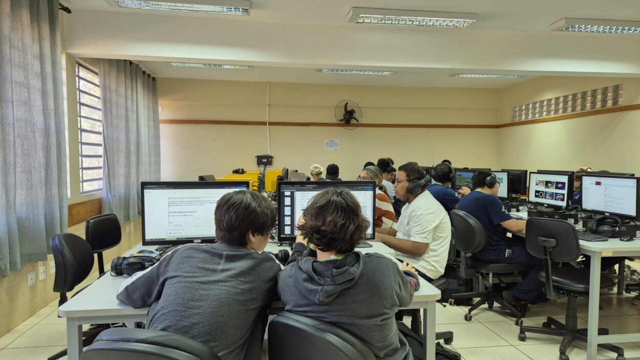 A primeira imagem mostra um grupo de estudantes em uma sala de aula. São aproximadamente 30 pessoas, sendo alunos e professora. O ambiente é uma sala de aula com mesas redondas, cadeiras azuis e um piso de madeira escura. Há um ar-condicionado fixado na parede ao fundo e luminárias no teto. Algumas mochilas e materiais escolares estão ao redor da sala. Na segunda fotografia há um grupo de seis pessoas, provavelmente estudantes e uma professora, posando sorridentes em frente a um painel que diz "Clube de Ciências - Capivara’s Tech". Eles estão sentados em bancos e mesas. Há pequenos vasos de planta como decoração. O grupo está em um pátio com dois tons de cinza e um cartaz adicional na parede ao lado do banner principal. Na terceira fotografia tem uma sala de informática com diversos alunos utilizando computadores. Os estudantes estão usando fones de ouvido e parecem concentrados nas telas, que exibem páginas da web, apresentações e imagens. Na frente da imagem, dois estudantes estão trabalhando juntos em um computador. As mesas e cadeiras são organizadas em fileiras, e cada estação possui um monitor, teclado e headset. A quarta fotografia mostra um grupo de estudantes junto a uma professora em uma sala de aula. São 12 pessoas no total. A professora, que está sorrindo, veste um suéter branco claro com cachecol verde escuro. O grupo está posando para a foto entre carteiras escolares organizadas em fileiras. Ao fundo, há um armário metálico. Na última foto, há um grupo de estudantes posando para uma foto com um professor em uma sala de aula. São 11 pessoas no total. A sala tem paredes de blocos de concreto pintadas de branco, com janelas verdes à direita, carteiras escolares organizadas e alguns ventiladores presos à parede. No fundo, também há um aparelho de ar-condicionado e iluminação fluorescente no teto.