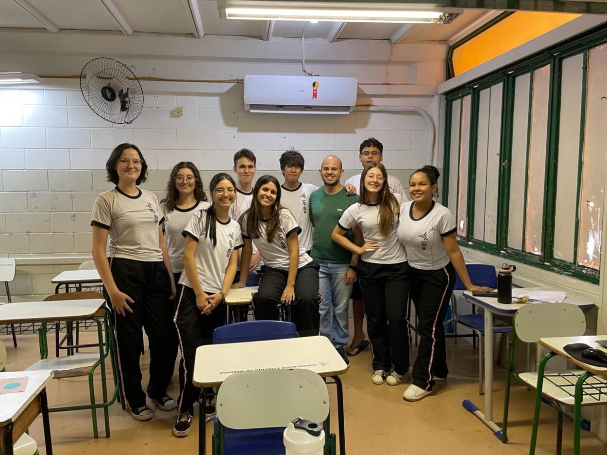 A primeira imagem mostra um grupo de estudantes em uma sala de aula. São aproximadamente 30 pessoas, sendo alunos e professora. O ambiente é uma sala de aula com mesas redondas, cadeiras azuis e um piso de madeira escura. Há um ar-condicionado fixado na parede ao fundo e luminárias no teto. Algumas mochilas e materiais escolares estão ao redor da sala. Na segunda fotografia há um grupo de seis pessoas, provavelmente estudantes e uma professora, posando sorridentes em frente a um painel que diz "Clube de Ciências - Capivara’s Tech". Eles estão sentados em bancos e mesas. Há pequenos vasos de planta como decoração. O grupo está em um pátio com dois tons de cinza e um cartaz adicional na parede ao lado do banner principal. Na terceira fotografia tem uma sala de informática com diversos alunos utilizando computadores. Os estudantes estão usando fones de ouvido e parecem concentrados nas telas, que exibem páginas da web, apresentações e imagens. Na frente da imagem, dois estudantes estão trabalhando juntos em um computador. As mesas e cadeiras são organizadas em fileiras, e cada estação possui um monitor, teclado e headset. A quarta fotografia mostra um grupo de estudantes junto a uma professora em uma sala de aula. São 12 pessoas no total. A professora, que está sorrindo, veste um suéter branco claro com cachecol verde escuro. O grupo está posando para a foto entre carteiras escolares organizadas em fileiras. Ao fundo, há um armário metálico. Na última foto, há um grupo de estudantes posando para uma foto com um professor em uma sala de aula. São 11 pessoas no total. A sala tem paredes de blocos de concreto pintadas de branco, com janelas verdes à direita, carteiras escolares organizadas e alguns ventiladores presos à parede. No fundo, também há um aparelho de ar-condicionado e iluminação fluorescente no teto.