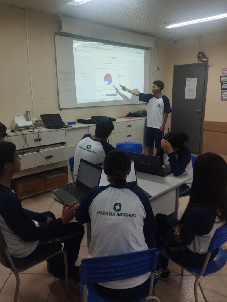 Na primeira imagem há uma sala de aula onde um grupo de estudantes está participando de uma atividade. Alguns estão sentados ao redor de uma mesa utilizando notebooks, enquanto um aluno em pé apresenta algo projetado em um quadro branco, apontando para um gráfico exibido na tela. O ambiente tem paredes claras, iluminação fluorescente e móveis escolares, como cadeiras e armários. Na segunda fotografia tem um grupo de estudantes reunidos em um ambiente escolar ao ar livre, em frente a um jardim vertical com diversas plantas verdes. Eles estão distribuídos em pé e agachados, posando para a foto. O local tem piso de tijolos, árvores, telhados de cerâmica e áreas cobertas ao fundo. A terceira imagem mostra um grupo de estudantes e um adulto reunidos em um espaço ao ar livre, posando para uma foto em frente a uma grande escultura de uma figura humana com os braços abertos. O grupo está distribuído sobre um pequeno morro coberto de grama, alguns sentados e outros em pé, enquanto ao fundo há muitas árvores, formando um cenário verde e natural. Na quarta imagem há um grupo de estudantes e dois adultos posando para uma foto em frente a uma placa de madeira com a palavra “Meliponário” e o desenho de uma abelha sorridente. Eles estão em um espaço externo com bastante vegetação, incluindo árvores altas e um caminho de pedras à esquerda. Ao fundo, vê-se um prédio de tijolos com telhado de cerâmica. Na quinta imagem tem um grupo de estudantes e alguns adultos reunidos para uma foto em um ambiente escolar externo. Eles estão em frente a uma parede branca com detalhes verdes e uma janela comprida com grades. O grupo está posicionado em duas fileiras, com alguns agachados na frente e outros em pé atrás, todos sorrindo e olhando para a câmera. A última fotografia mostra um grupo grande de pessoas, formado por adolescentes e um adulto, posando para uma foto em uma sala. Eles estão organizados em duas fileiras: alguns sentados ou agachados na frente e outros em pé atrás. A sala tem paredes claras, ventiladores fixados na parede e um quadro branco ao fundo. Há cartazes e avisos na parede. À esquerda, há uma mesa com uma toalha vermelha e alguns objetos decorativos para um evento escolar.