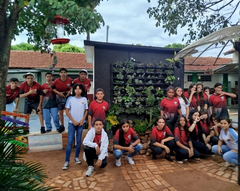 Na primeira imagem há uma sala de aula onde um grupo de estudantes está participando de uma atividade. Alguns estão sentados ao redor de uma mesa utilizando notebooks, enquanto um aluno em pé apresenta algo projetado em um quadro branco, apontando para um gráfico exibido na tela. O ambiente tem paredes claras, iluminação fluorescente e móveis escolares, como cadeiras e armários. Na segunda fotografia tem um grupo de estudantes reunidos em um ambiente escolar ao ar livre, em frente a um jardim vertical com diversas plantas verdes. Eles estão distribuídos em pé e agachados, posando para a foto. O local tem piso de tijolos, árvores, telhados de cerâmica e áreas cobertas ao fundo. A terceira imagem mostra um grupo de estudantes e um adulto reunidos em um espaço ao ar livre, posando para uma foto em frente a uma grande escultura de uma figura humana com os braços abertos. O grupo está distribuído sobre um pequeno morro coberto de grama, alguns sentados e outros em pé, enquanto ao fundo há muitas árvores, formando um cenário verde e natural. Na quarta imagem há um grupo de estudantes e dois adultos posando para uma foto em frente a uma placa de madeira com a palavra “Meliponário” e o desenho de uma abelha sorridente. Eles estão em um espaço externo com bastante vegetação, incluindo árvores altas e um caminho de pedras à esquerda. Ao fundo, vê-se um prédio de tijolos com telhado de cerâmica. Na quinta imagem tem um grupo de estudantes e alguns adultos reunidos para uma foto em um ambiente escolar externo. Eles estão em frente a uma parede branca com detalhes verdes e uma janela comprida com grades. O grupo está posicionado em duas fileiras, com alguns agachados na frente e outros em pé atrás, todos sorrindo e olhando para a câmera. A última fotografia mostra um grupo grande de pessoas, formado por adolescentes e um adulto, posando para uma foto em uma sala. Eles estão organizados em duas fileiras: alguns sentados ou agachados na frente e outros em pé atrás. A sala tem paredes claras, ventiladores fixados na parede e um quadro branco ao fundo. Há cartazes e avisos na parede. À esquerda, há uma mesa com uma toalha vermelha e alguns objetos decorativos para um evento escolar.